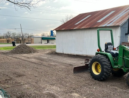 Green tractor grading a gravel drive in front of a white shop building with a patinaed metal roof.