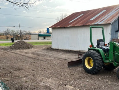 Green tractor grading a gravel drive in front of a white shop building with a patinaed metal roof.