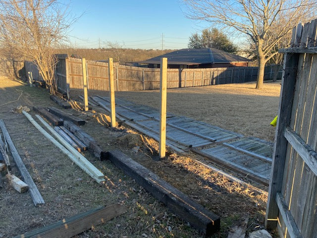 Wooden framing materials on the ground with a wooden fence and trees in the background while post are being replaced during picuture