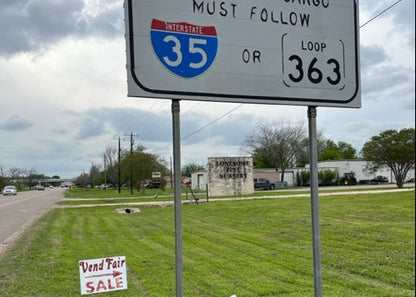 Road sign indicating oversized cargo must follow Interstate 35 or Loop 363 with a grassy area, road, and a neighboring business in the background.